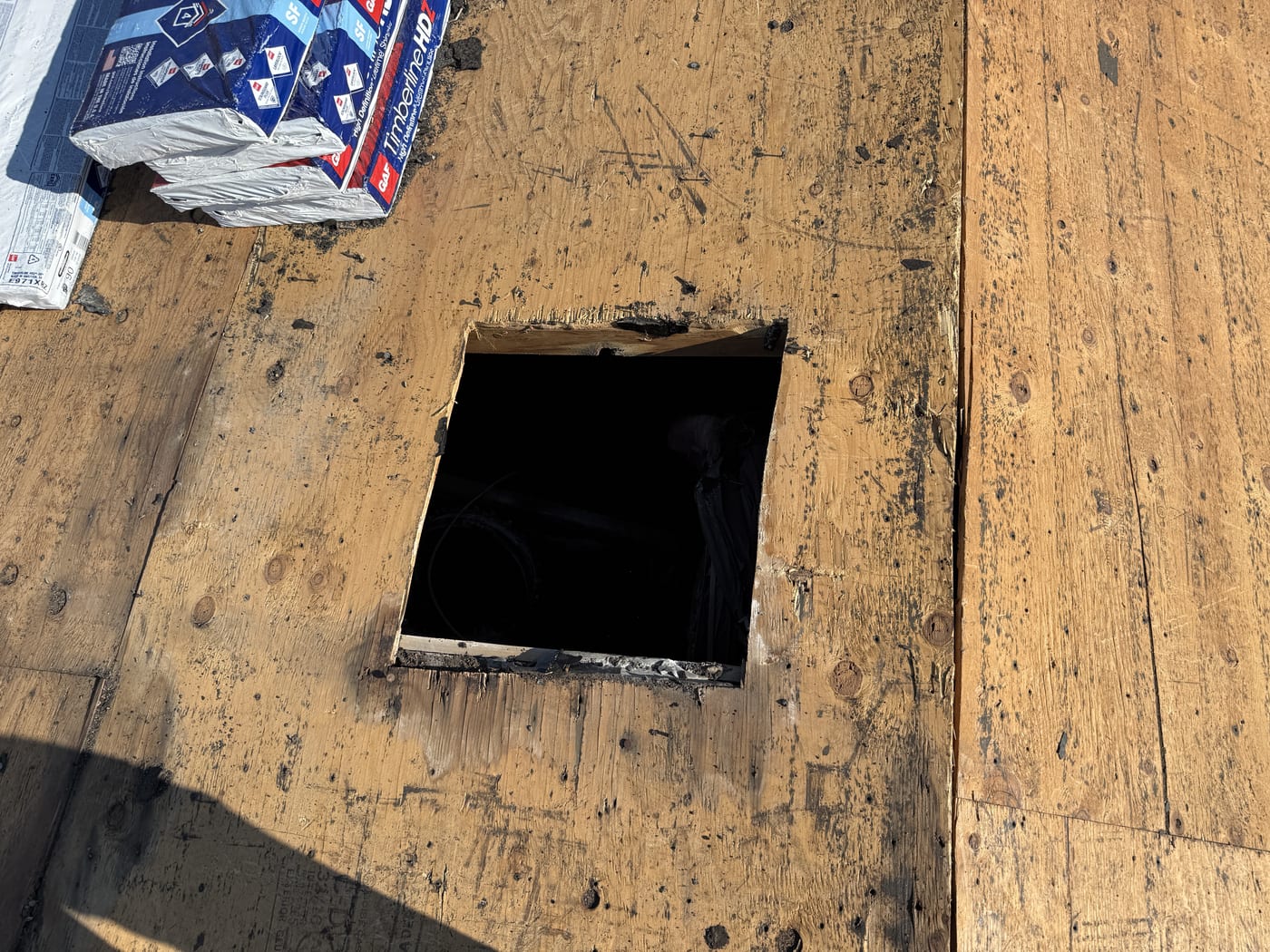 Close-up of square penetration hole cut in bare OSB roof deck, GAF shingle packages visible at edge