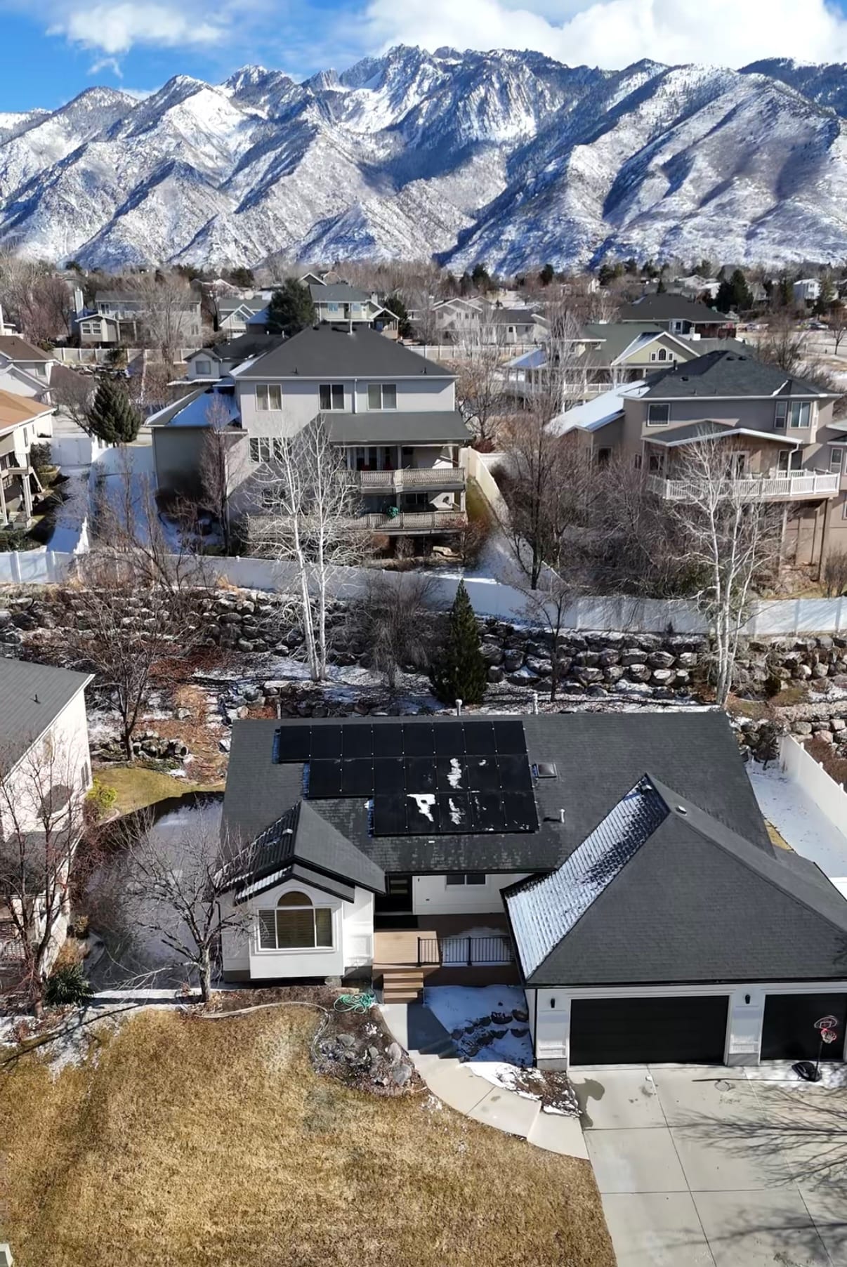 [black-on-black Trina panels] High drone aerial of residential neighborhood in winter showing house with rooftop solar, snow-covered yards, and dramatic Wasatch mountain backdrop