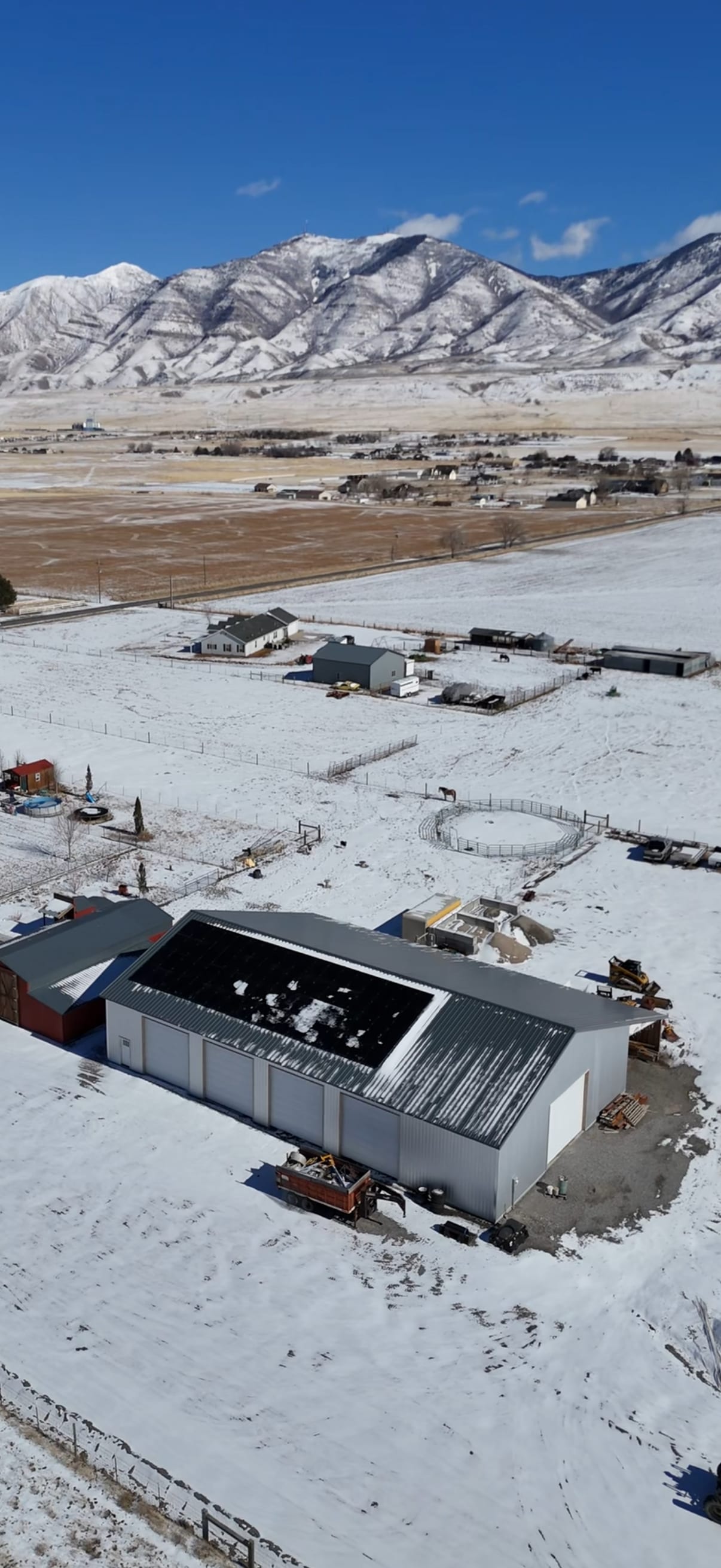 [Tier 1 Trina panels] Wide vertical drone aerial of rural property showing metal barn with rooftop solar, surrounding outbuildings, snow-covered fields, and mountain backdrop