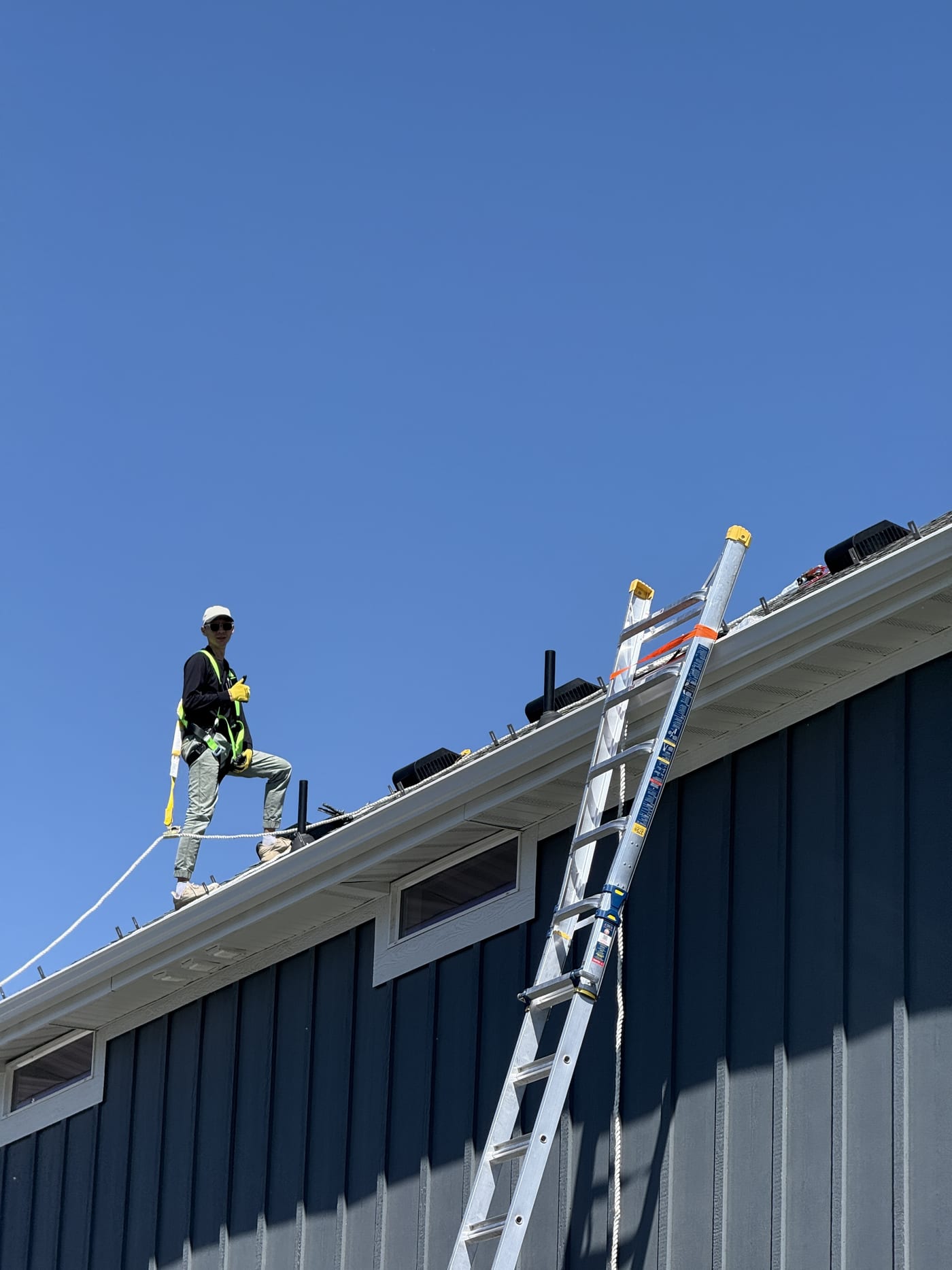 Worker climbing ladder on blue house with safety harness, clear sky