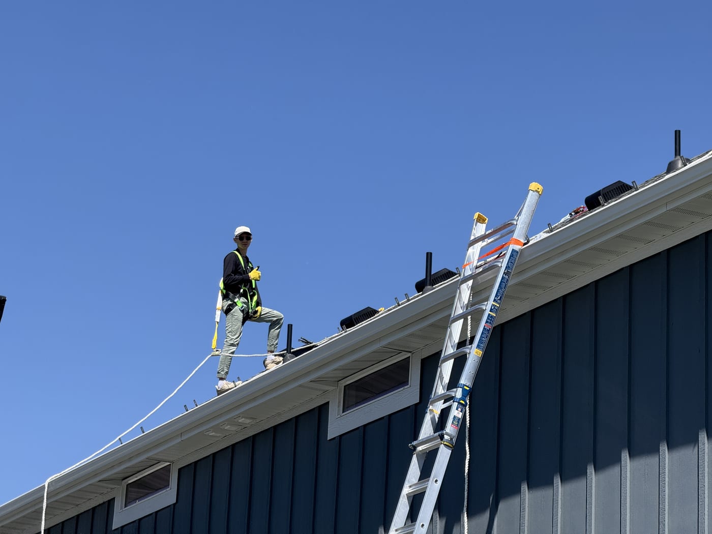 Worker on roof edge with ladder against blue siding, blue sky