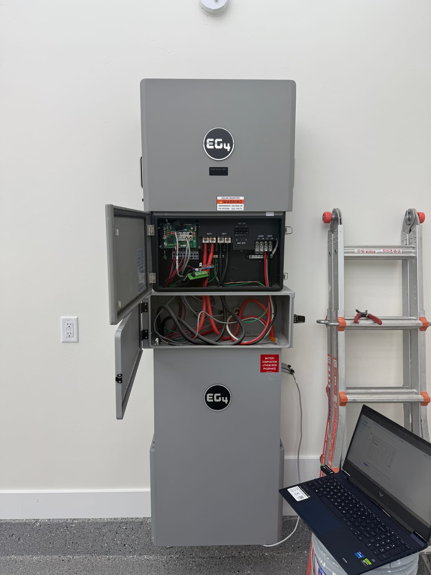 EG4 GridBOSS open enclosure showing internal busbars, breakers, and DC/AC wiring during commissioning, laptop on top for setup, ladder propped behind on white garage wall