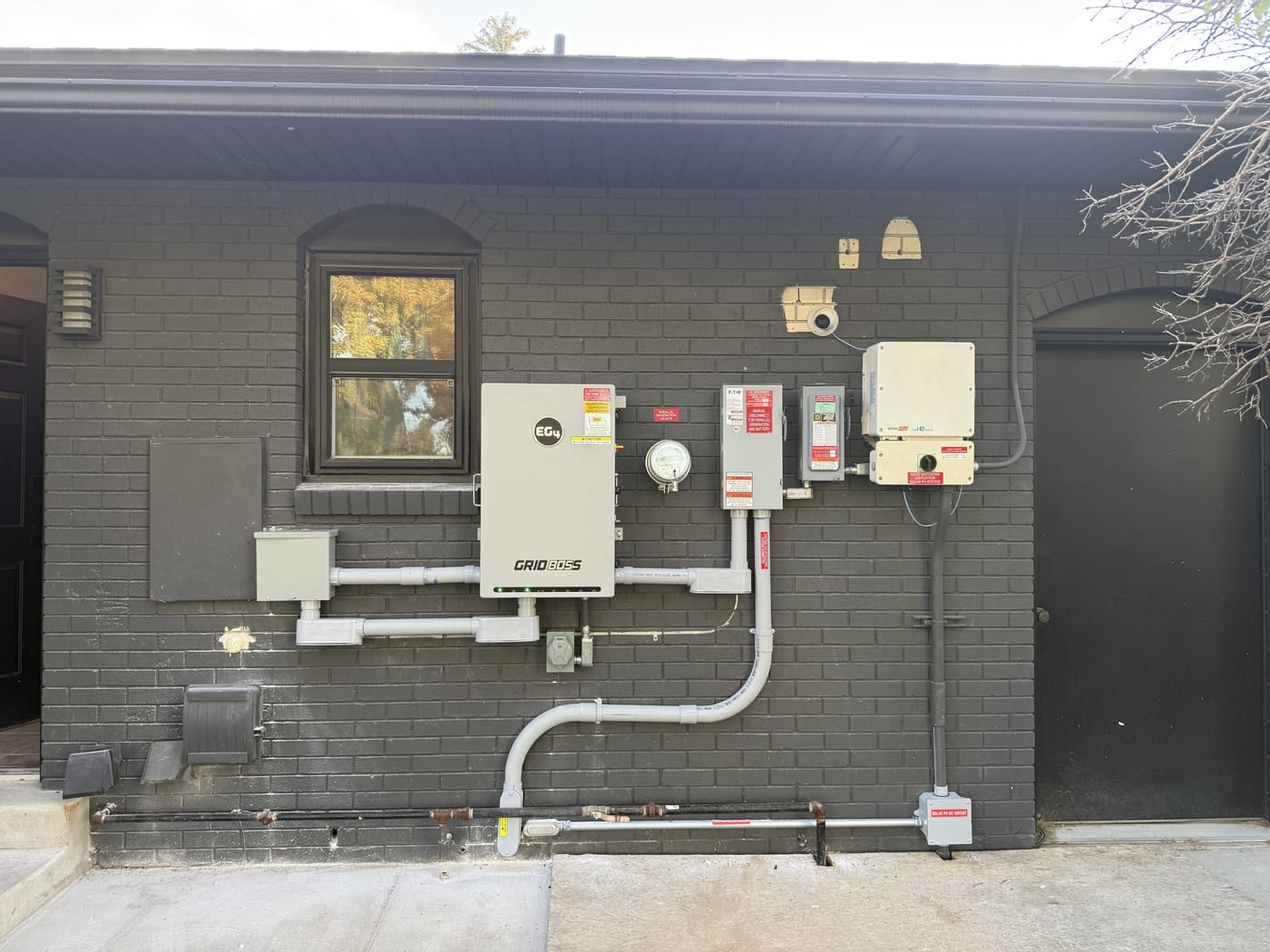 Wide view of EG4 GridBOSS inverter system with meter and disconnects on dark brick exterior wall