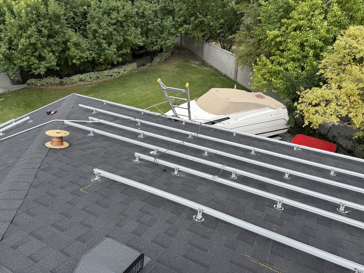 Racking rails installed on dark shingle roof with ladder and green yard visible below