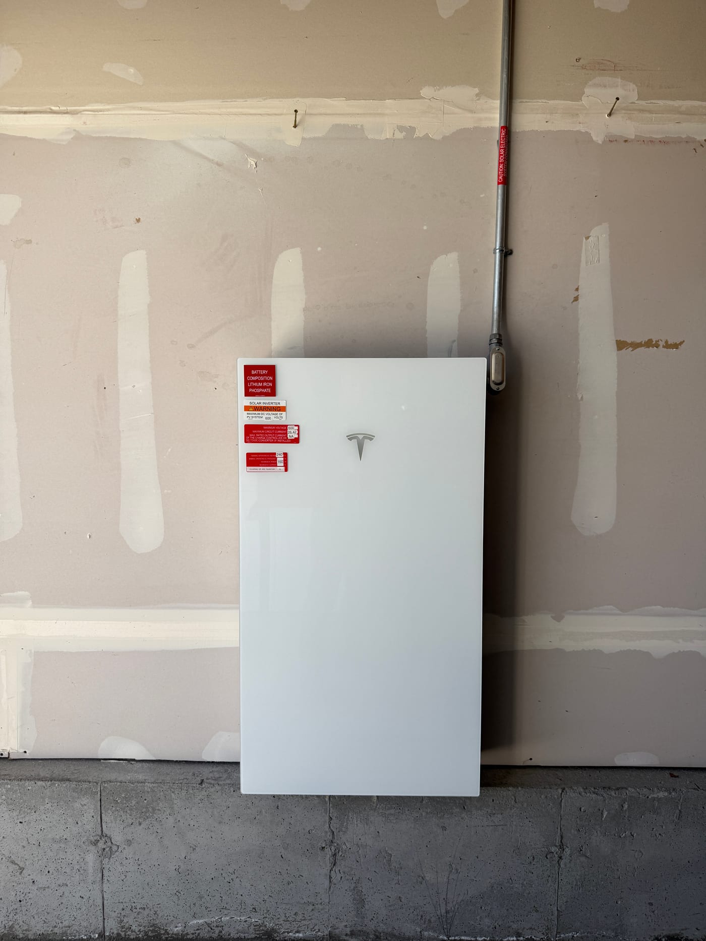 Tesla Powerwall 3 mounted on unfinished drywall wall in garage with red rapid-shutdown and warning stickers, conduit running across wall, install in progress