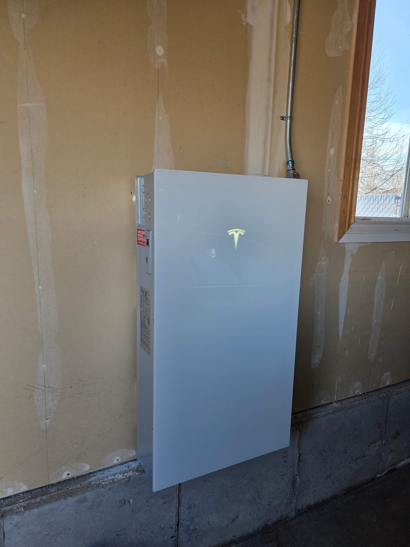 Tesla Powerwall 3 with illuminated Tesla logo mounted on unfinished garage wall with conduit running to ceiling