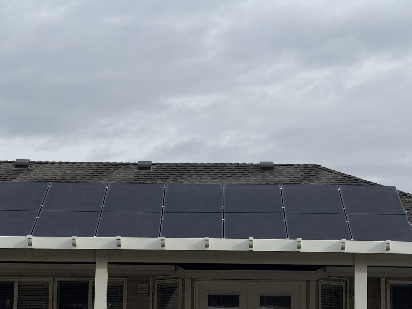 [black-on-black panels] Close-up looking up at solar panel row on shingle roof showing racking clamps and panel undersides, overcast sky (image rotated)