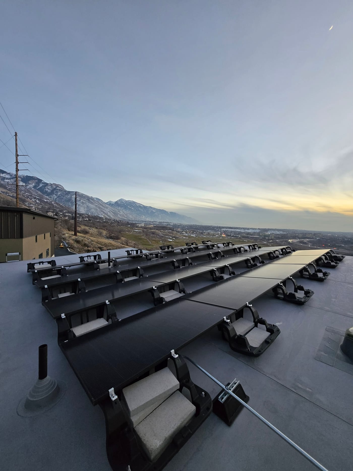 Multiple rows of ballasted solar panels on flat roof with snow-capped mountains and valley view, dramatic composition