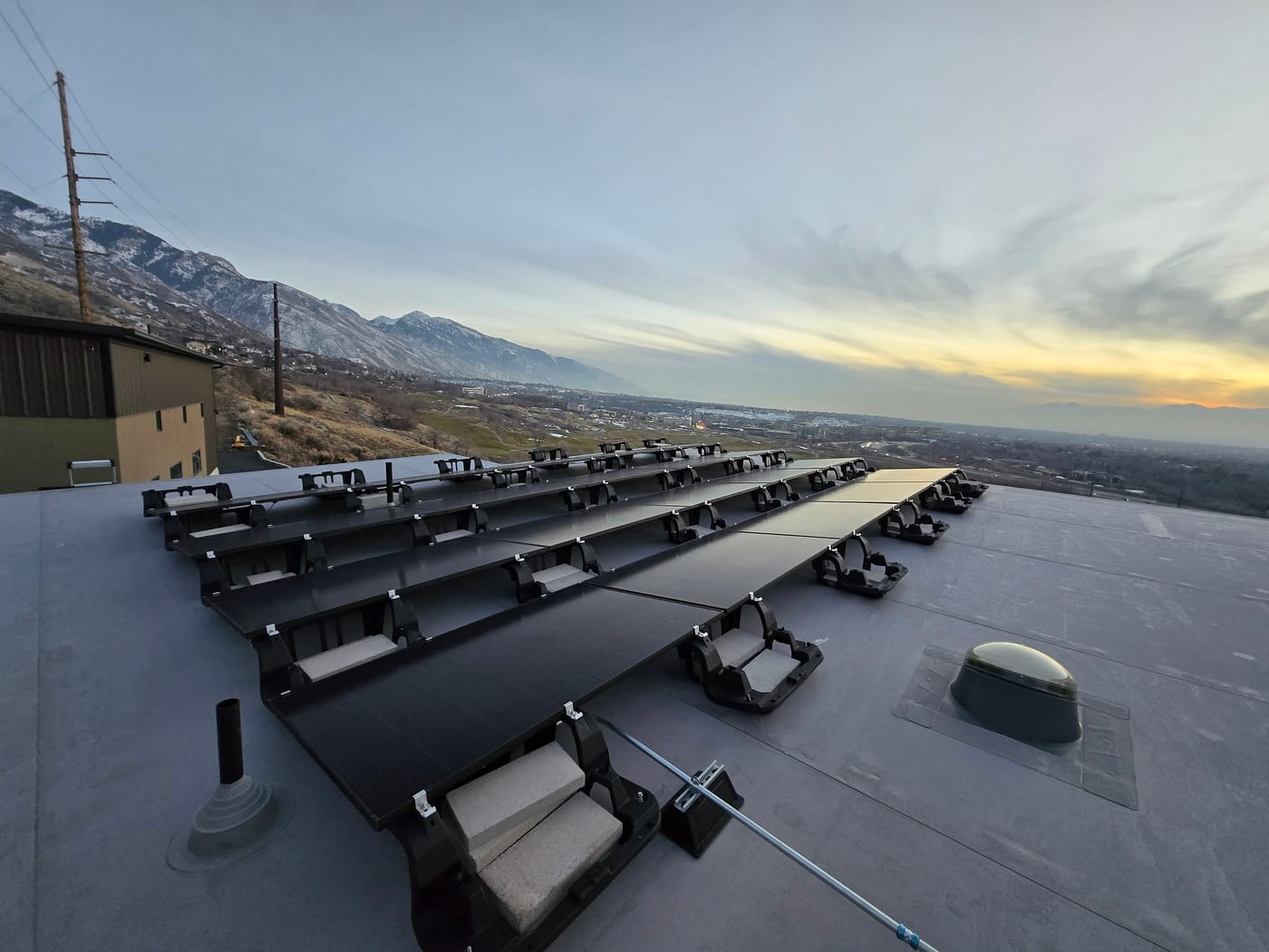 Rows of ballasted solar panels on flat roof facing snow-capped Wasatch mountains, outbuilding visible