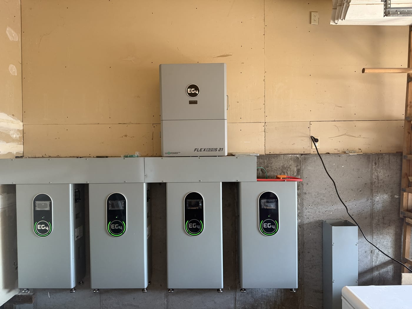 Five EG4 batteries in a row with FlexBOSS 21 inverter above on plywood backboard, all green LEDs active, garage setting