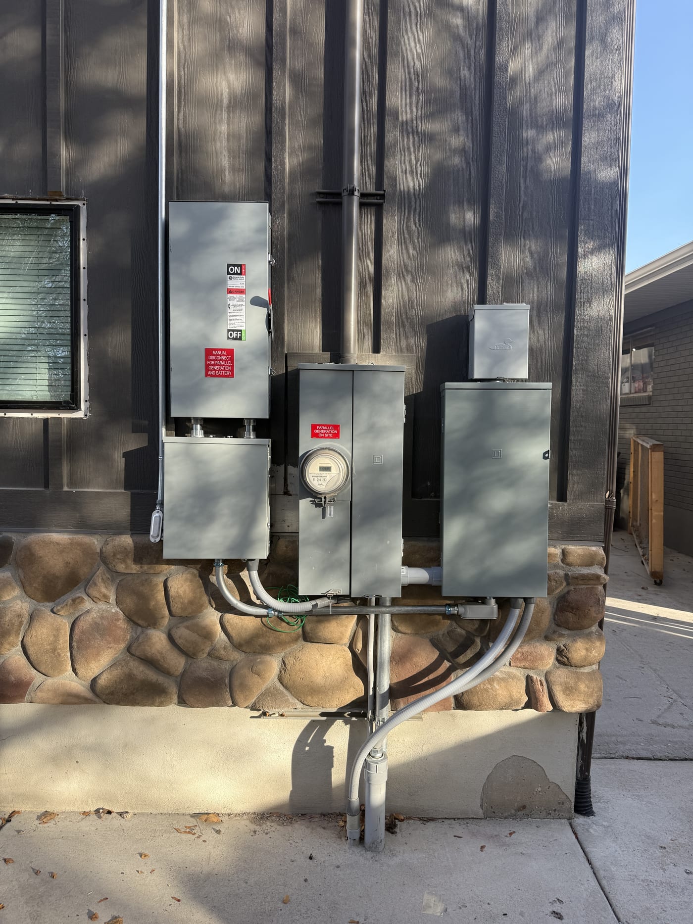 Side of house showing electrical equipment array including meter, electrical panels, disconnects, and conduit runs on stone and stucco wall