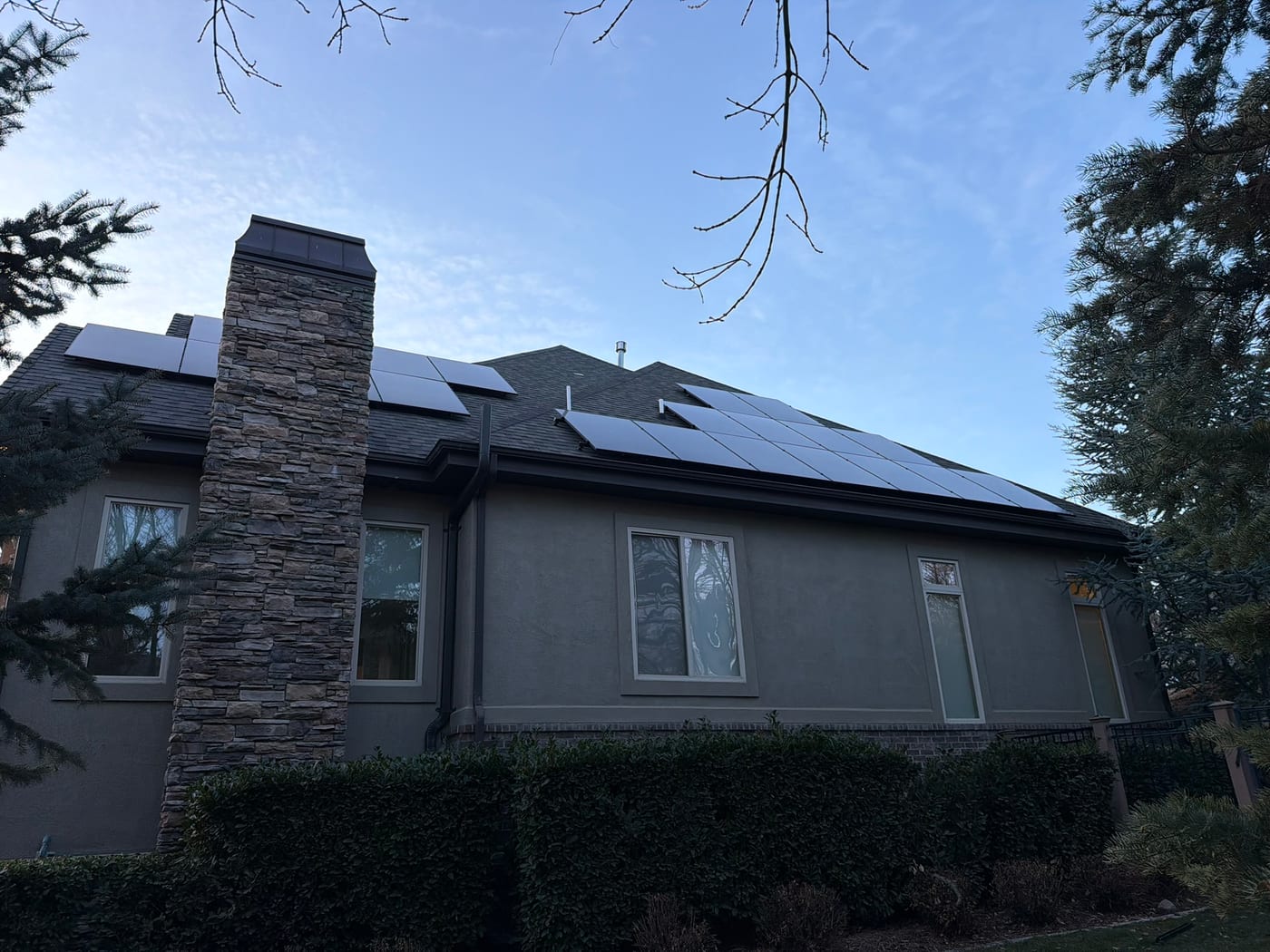 Rear view of dark stucco home with stone chimney showing solar panels on both roof faces, mature landscaping and blue sky