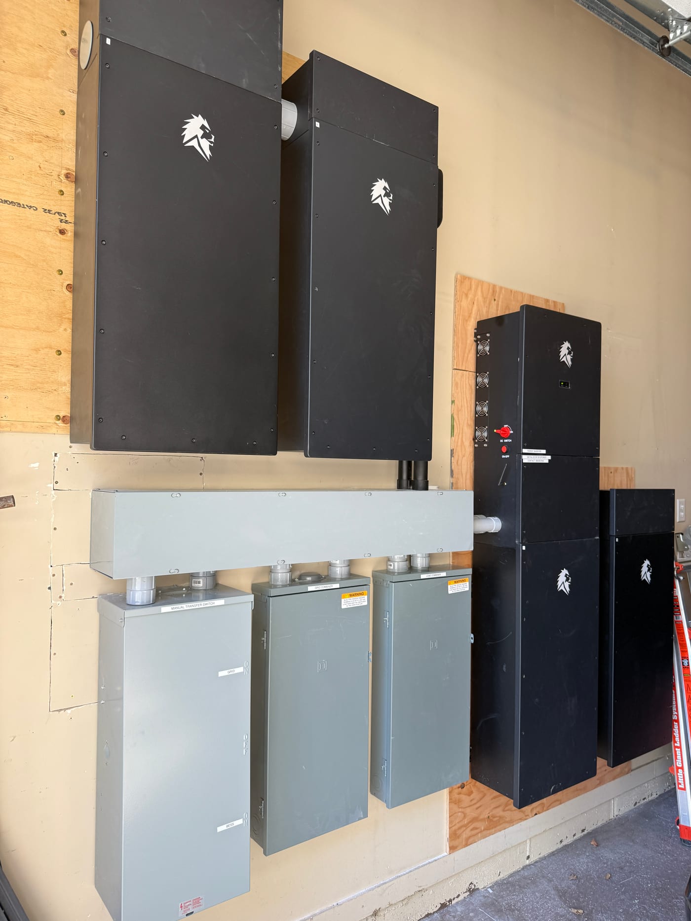 Lion Energy battery system components staged on garage floor before mounting — black battery cabinets with lion logos and three gray Lion Energy disconnect/breaker panels arranged on plywood, mid-install